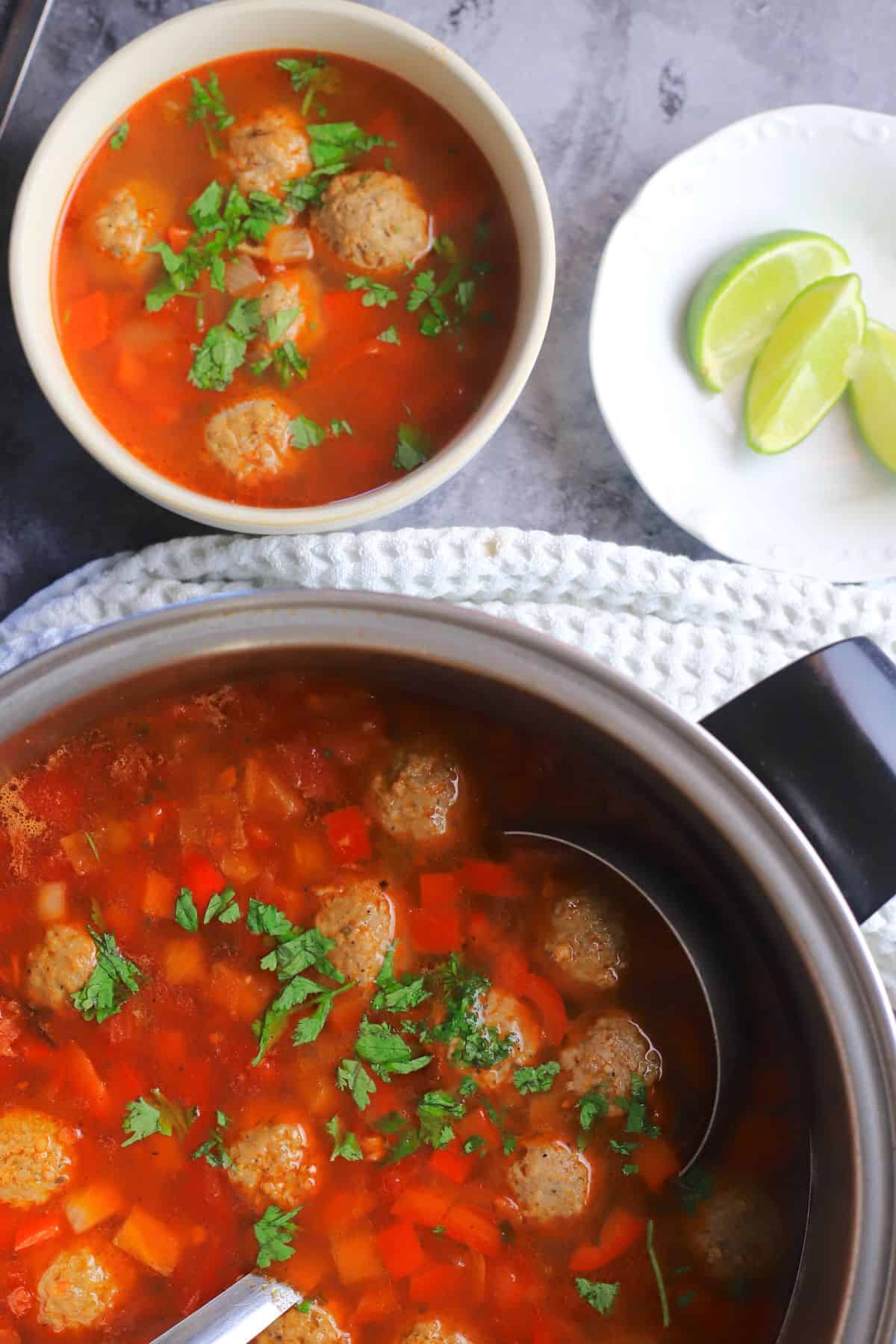 Low Carb Albondigas Mexican Meatball Soup Recipe in a bowl next to a white hand towel, pot of soup and lime wedges.