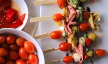 Low carb antipasto skewers on a platter next to a bowl of cherry tomatoes and roasted red peppers.