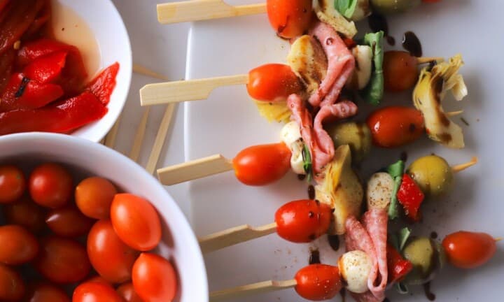 Low carb antipasto skewers on a platter next to a bowl of cherry tomatoes and roasted red peppers.