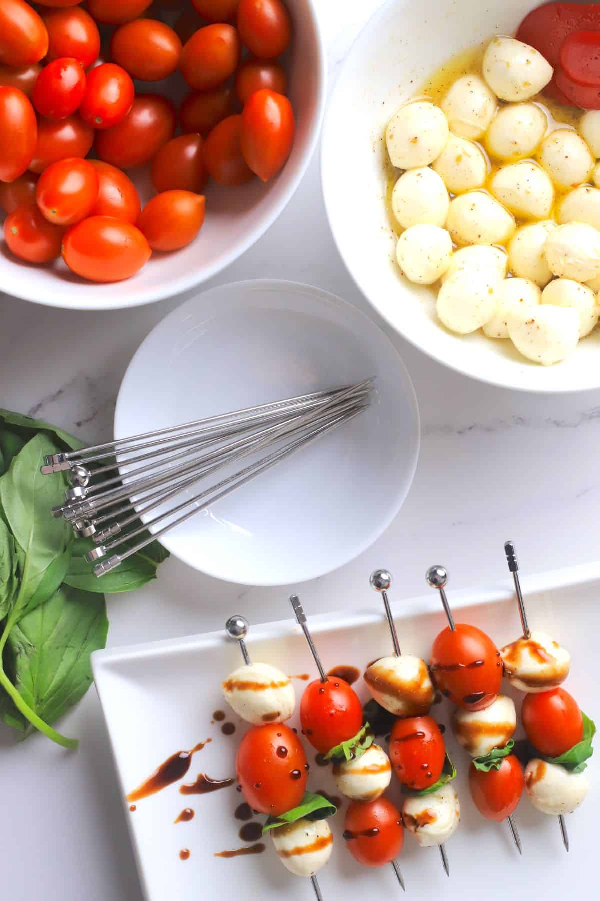 Overhead view of the the low carb caprese skewers on a platter next to a plate with skewers, leaves of basil, bowl of cherry tomatoes and a bowl of the marinated bocconcini.