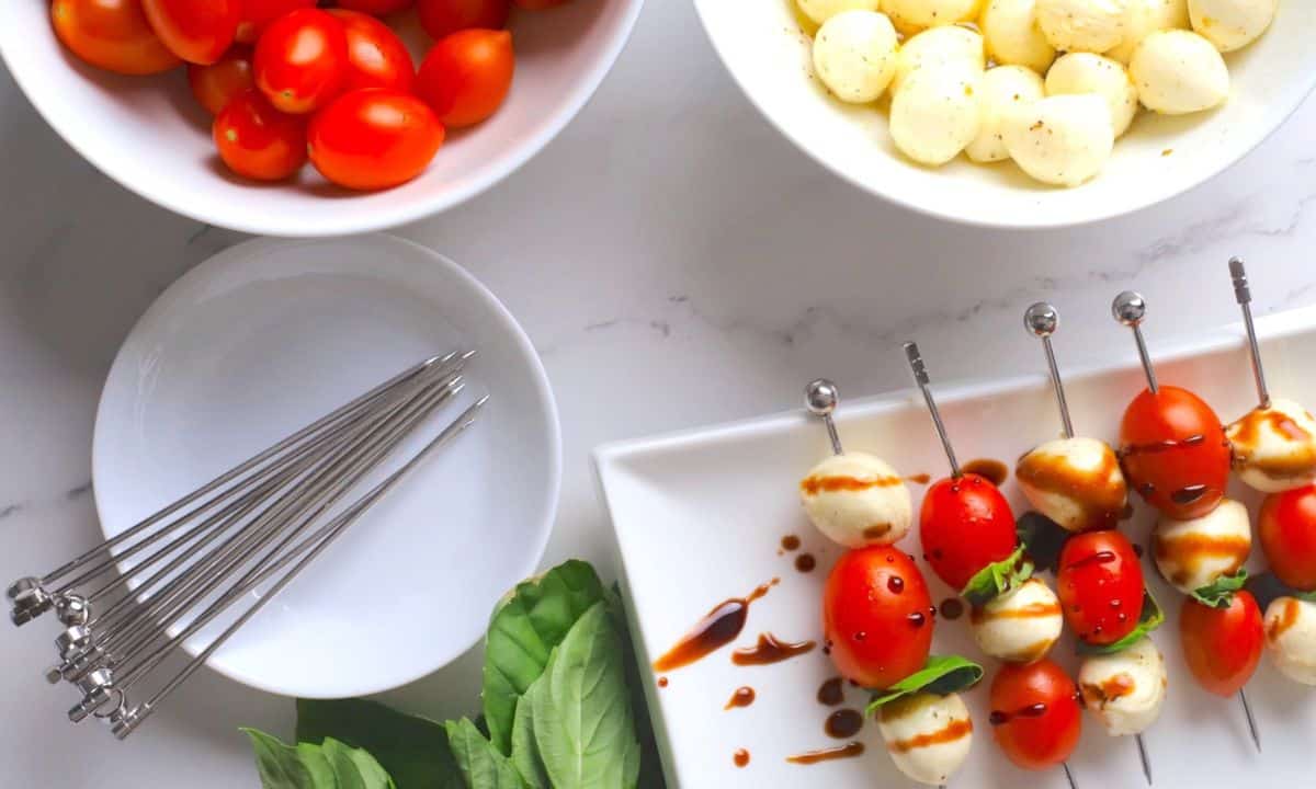 Overhead view of the the low carb caprese skewers on a platter next to a plate with skewers, leaves of basil, bowl of cherry tomatoes and a bowl of the marinated bocconcini.