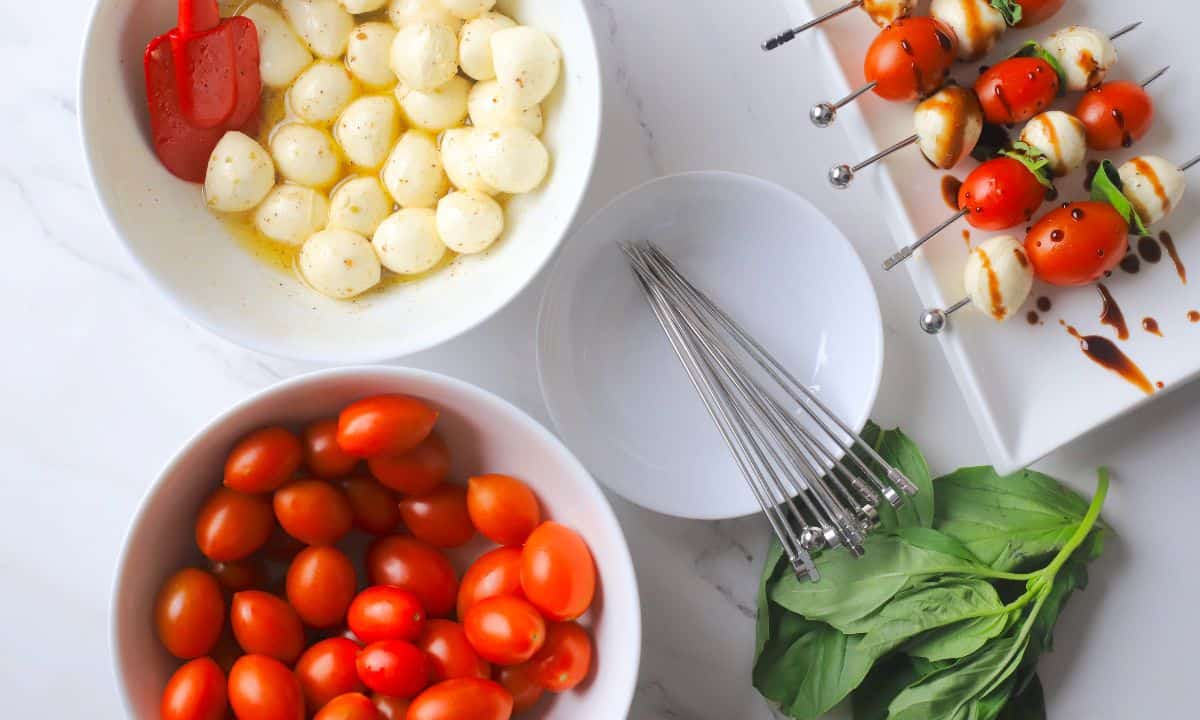 Overhead view of the the low carb caprese skewers on a platter next to a plate with skewers, leaves of basil, bowl of cherry tomatoes and a bowl of the marinated bocconcini.