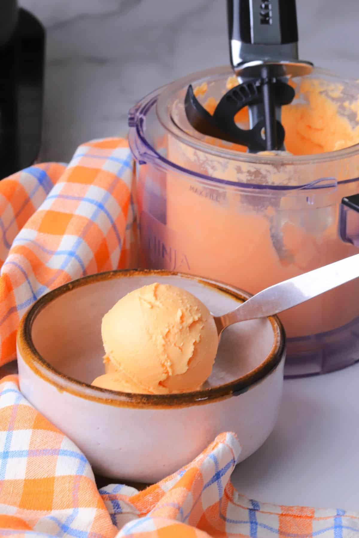 A bowl of sugar free orange creamsicle ice cream next to a creami pint with an ice cream scoop.