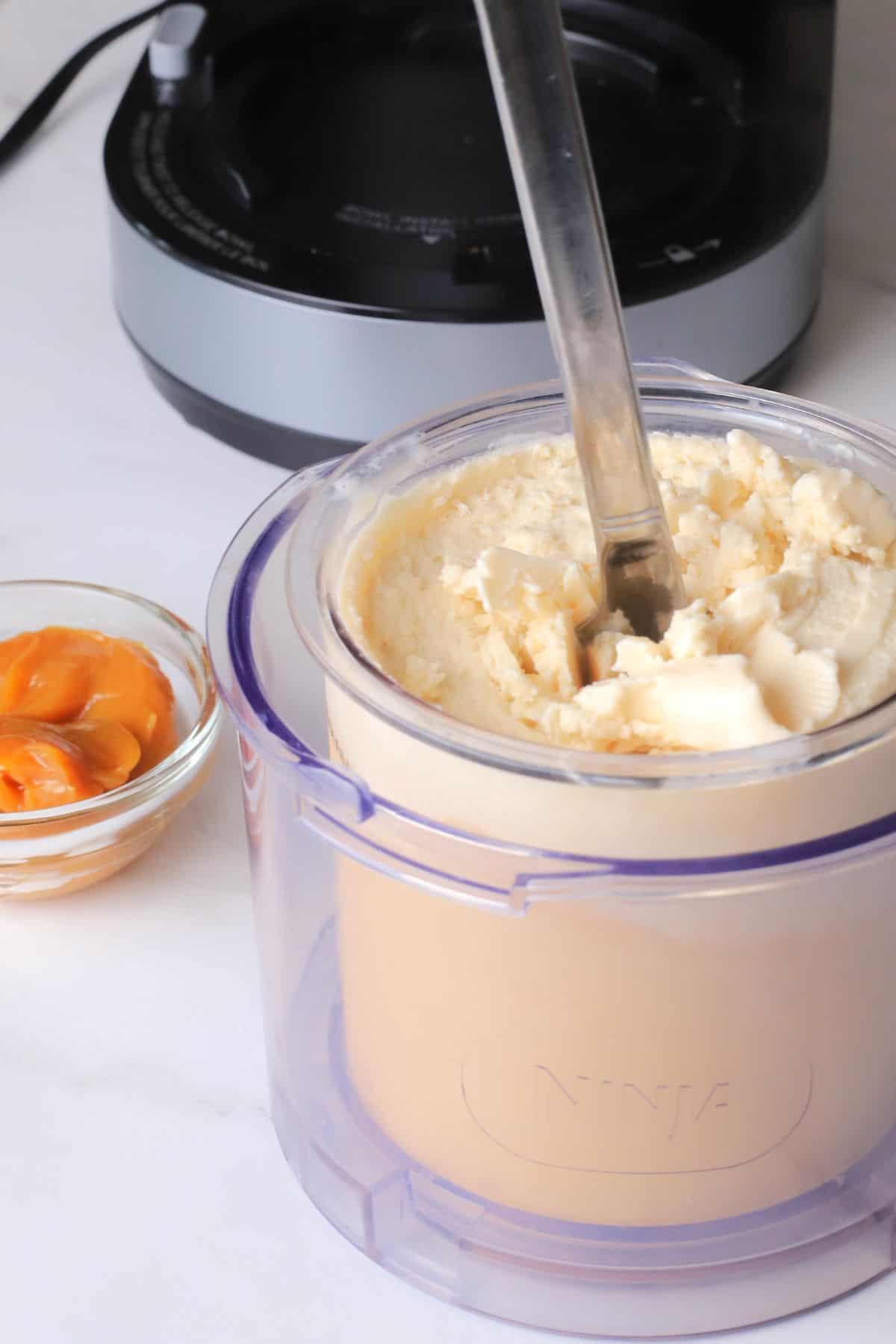 A knife in the centre of the ice cream in the creami pint next to a small bowl of caramel sauce.