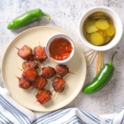 A plate with bacon wrapped pickles next to a side of ketchup, pickle slices, toothpicks and a jalapeno.