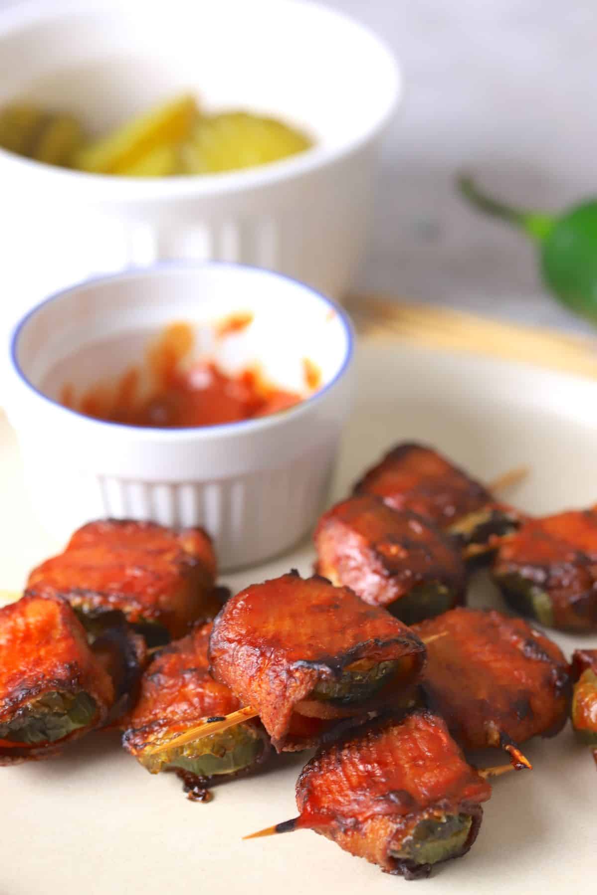 A plate with bacon wrapped pickles next to a side of ketchup, pickle slices, and a jalapeno.