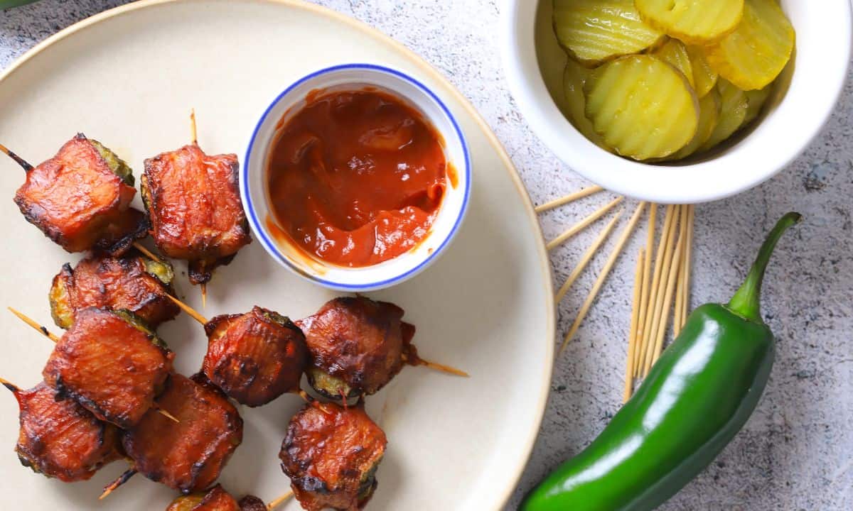 A plate with bacon wrapped pickles next to a side of ketchup, pickle slices, toothpicks and a jalapeno.