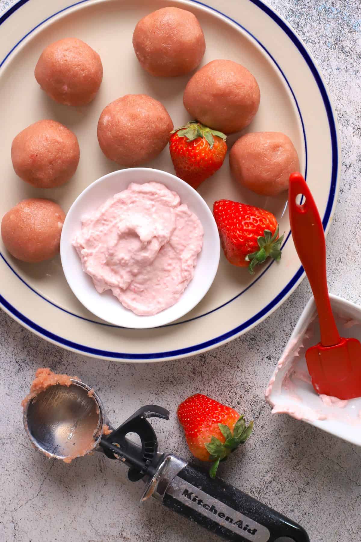 Strawberry protein bliss balls on a plate with a side of strawberry cream dip and fresh strawberries and an ice cream scoop.