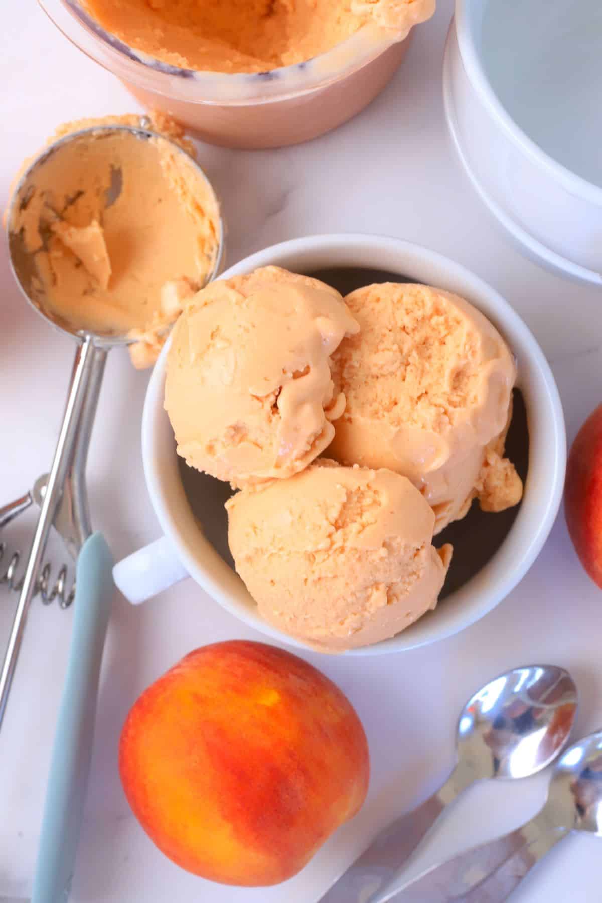 Bowl full of peach ice cream next to a peach, ice cream scoop and some spoons.