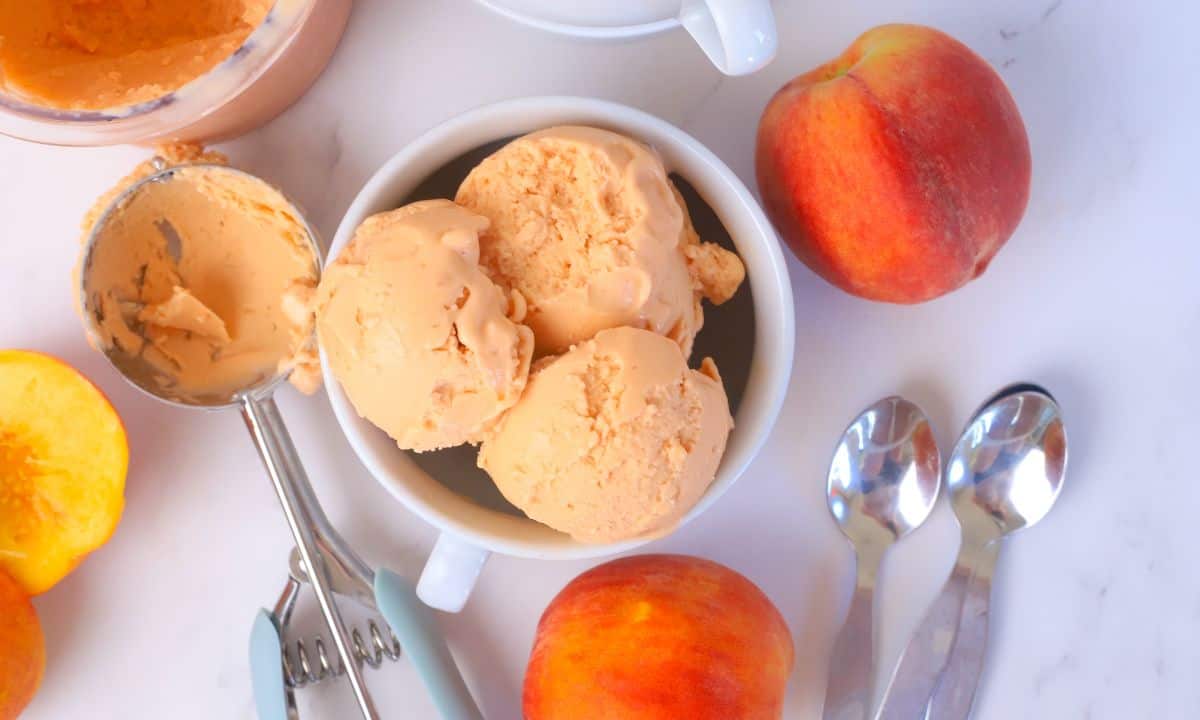 Bowl full of peach ice cream next to a peach, ice cream scoop and some spoons.