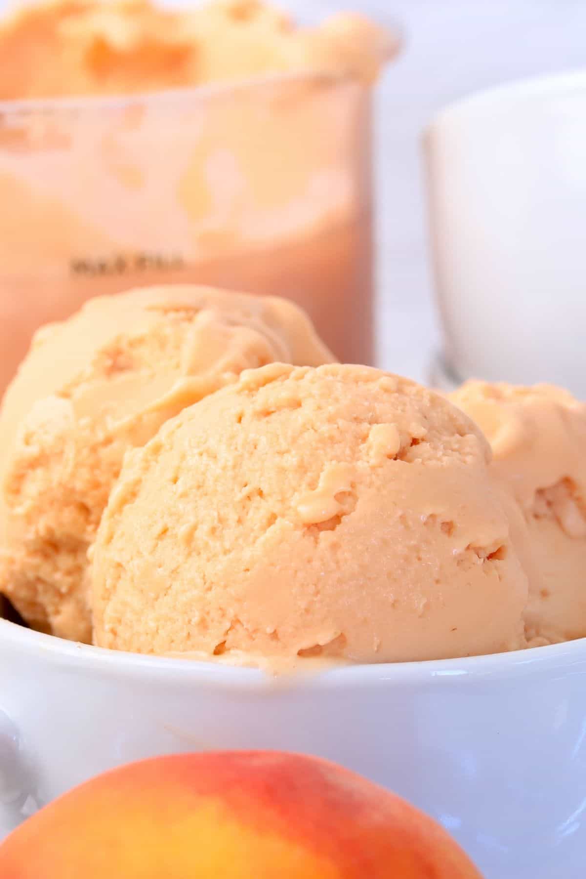 Bowl full of peach ice cream next to a peach, ice cream scoop and some spoons.