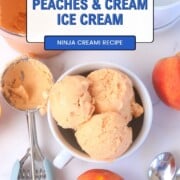 Bowl full of peach ice cream next to a peach, ice cream scoop and some spoons.