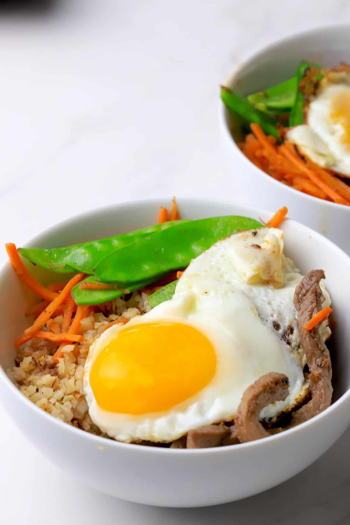 A white bowl filled with a bibimbap recipe of crispy cauliflower rice, beef, shredded carrots, snow peas and topped with a fried egg.