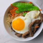 A white bowl filled with a bibimbap recipe of crispy cauliflower rice, beef, shredded carrots, snow peas and topped with a fried egg.
