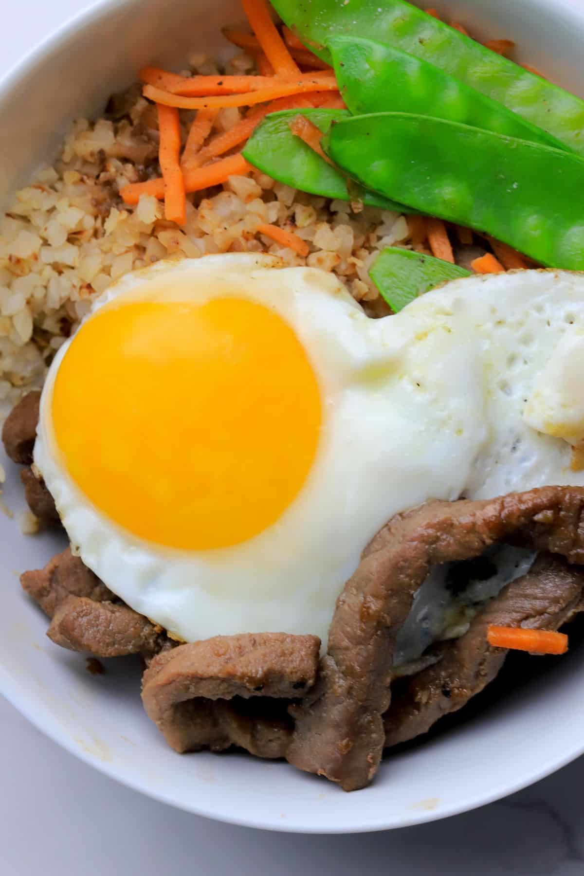 A white bowl filled with a bibimbap recipe of crispy cauliflower rice, beef, shredded carrots, snow peas and topped with a fried egg.