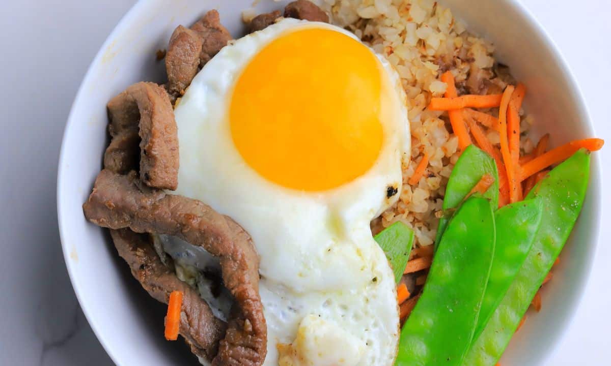 A white bowl filled with a bibimbap recipe of crispy cauliflower rice, beef, shredded carrots, snow peas and topped with a fried egg.