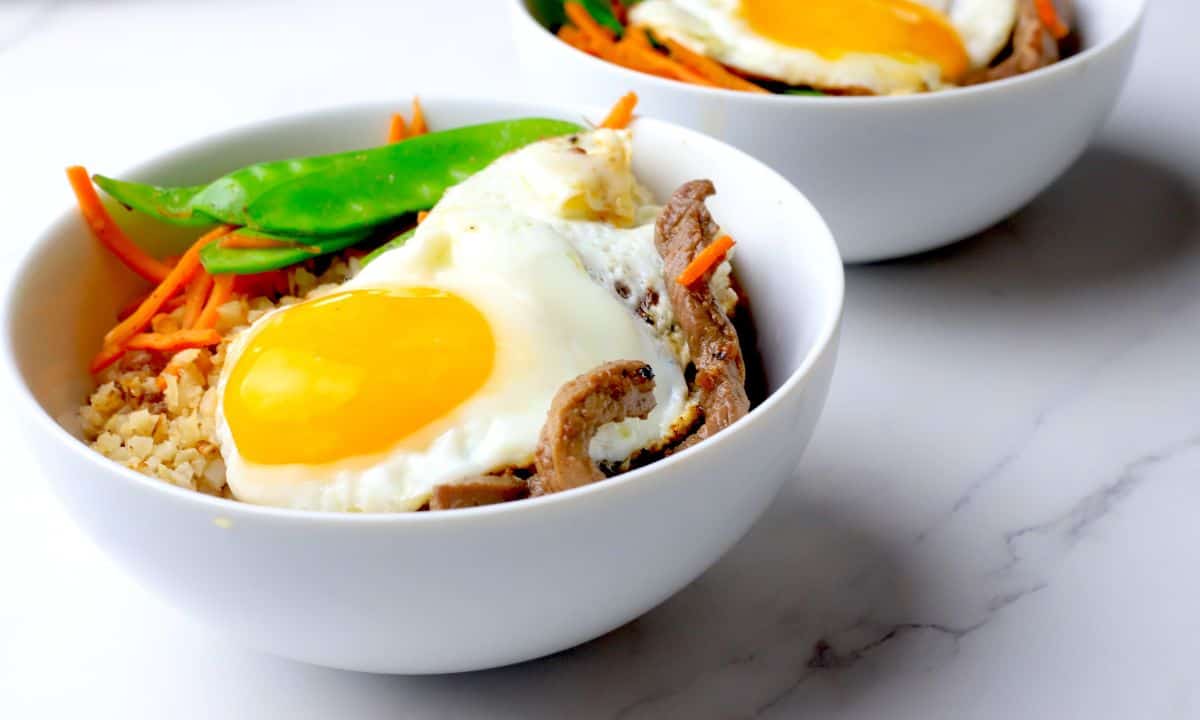A white bowl filled with a bibimbap recipe of crispy cauliflower rice, beef, shredded carrots, snow peas and topped with a fried egg.