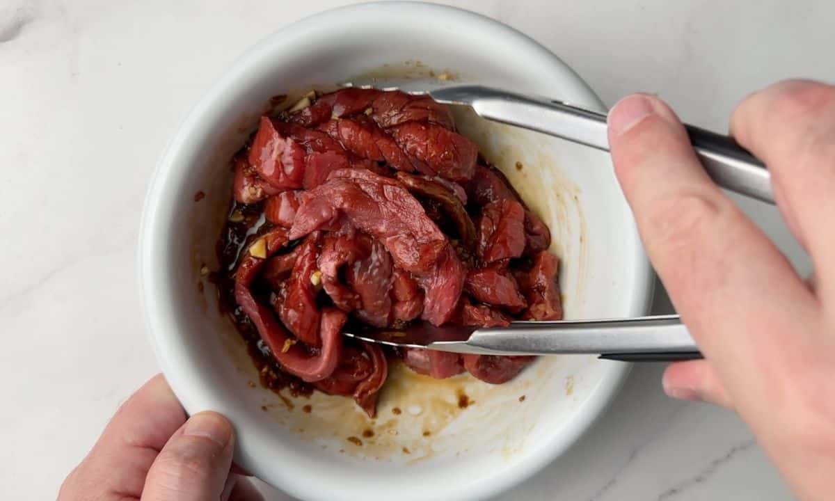 Adding the strips of beef to the marinade with tongs.