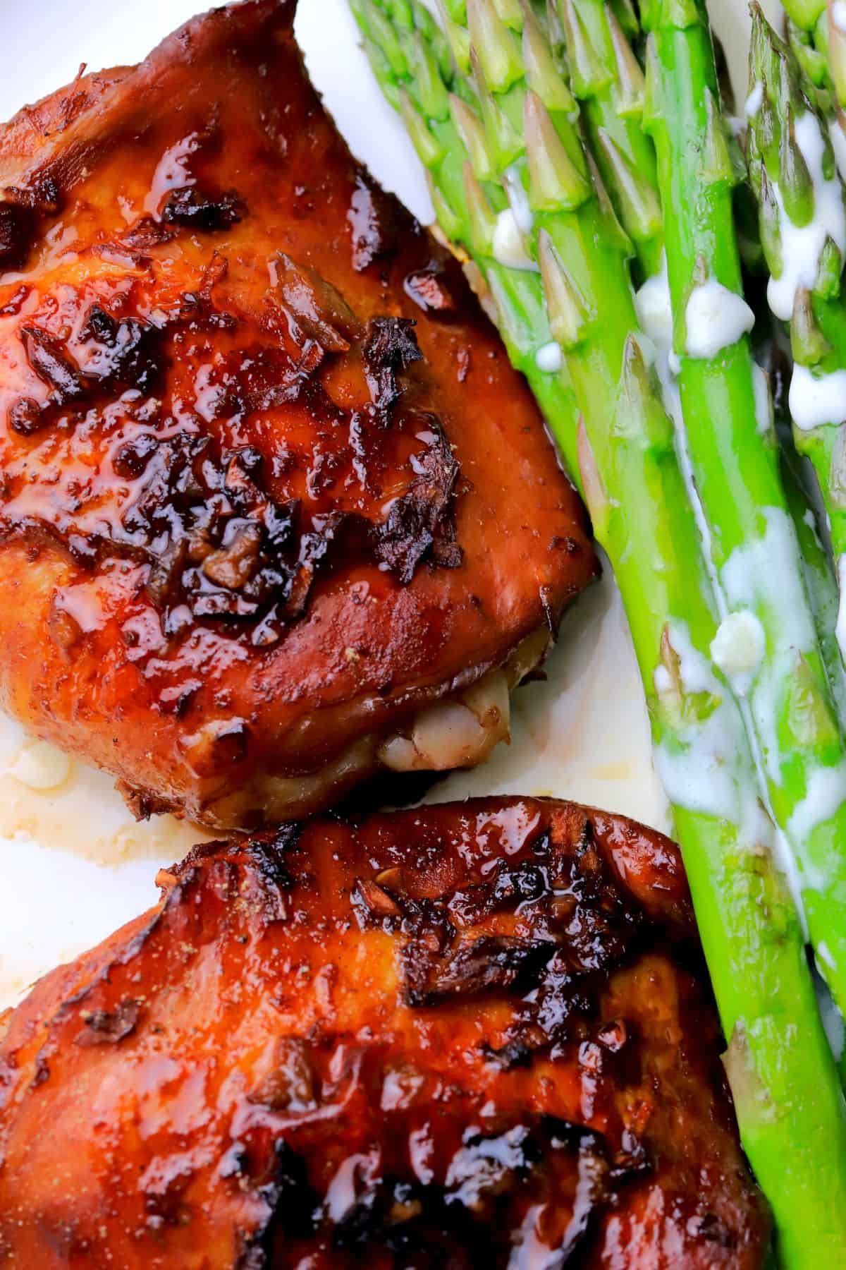 Two sticky chicken thighs with a low carb sauce on a plate next to asparagus spears.