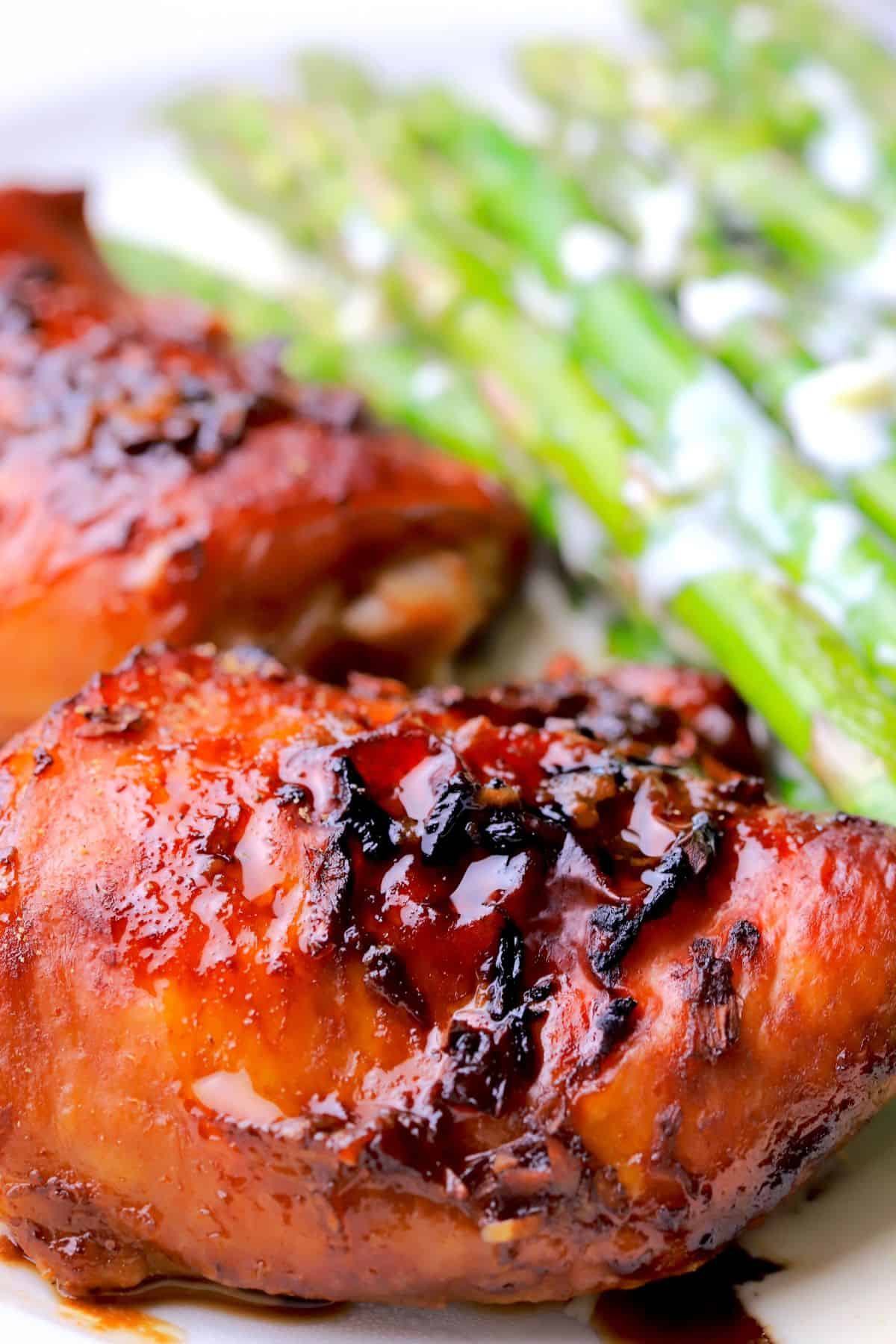 Two sticky chicken thighs with a low carb sauce on a plate next to asparagus spears.
