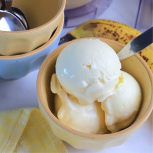 A bowl of sugar free banana ice cream next to small bowls, a banana and the ninja creami container.