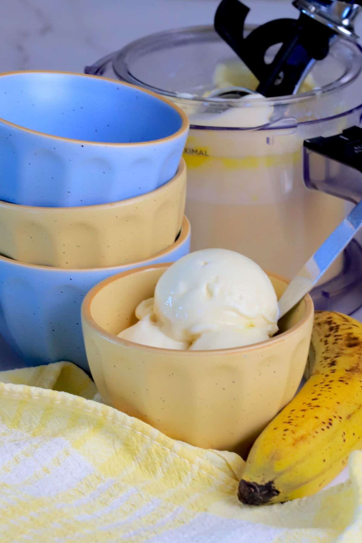 A bowl of sugar free banana ice cream next to small bowls, a banana and the ninja creami container.