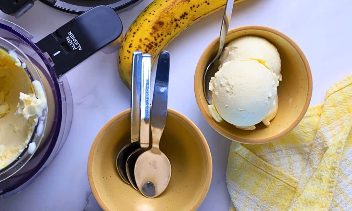 A bowl of sugar free banana ice cream next to small bowls, a banana and the ninja creami container.