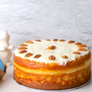 Sugar free carrot cake decorated with cream cheese frosting next to a coffee mug, a ceramic bunny rabbit and colored easter eggs.