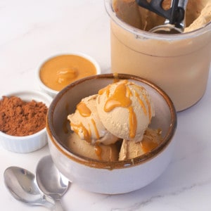 Sugar free chocolate peanut butter ice cream in a bowl topped with a drizzle of peanut butter with an ice cream scoop on the side and small bowls of chocolate protein powder and a small bowl of creamy peanut butter.
