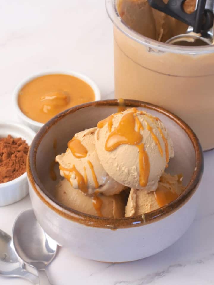Sugar free chocolate peanut butter ice cream in a bowl topped with a drizzle of peanut butter with an ice cream scoop on the side and small bowls of chocolate protein powder and a small bowl of creamy peanut butter.