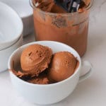 Sugar free triple chocolate ice cream in a white bowl next to the ninja creami pint and an ice cream scoop.