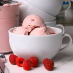 Ninja creami blue raspberry ice cream in a white bowl with raspberries and the ninja creami pint and more bowls in the background.
