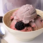 A bowl with scoops of ninja creami cherry blackberry ice cream with some fresh blackberries and spoons on the side with the ninja creami container in the background.