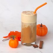 Wide shot of the Sugar-Free Iced Pumpkin Cream Chai Latte in a tall glass with an orange straw and a pumpkin, star anise and cinnamon stick next to the glass.