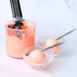 Sugar free watermelon ice cream in two glass bowls with spoons and the ninja creami pint next to them with an ice cream scoop,
