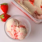 Strawberry cheesecake ice cream in a bowl next to strawberries, and another container with more ice cream and an ice cream scoop.