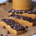Sugar free protein bars on brown parchment paper side by side with some chocolate chips and a jar of peanut butter in the background.