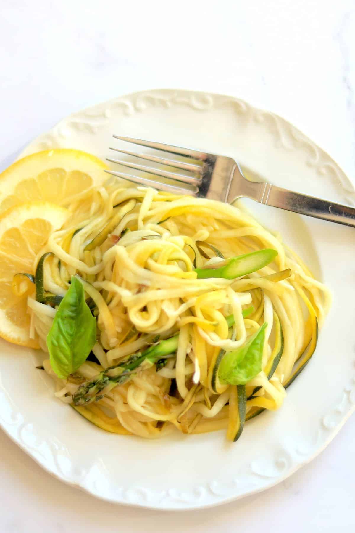 Zucchini veggie pasta on a white plate garnished with lemon slices and fresh basil.