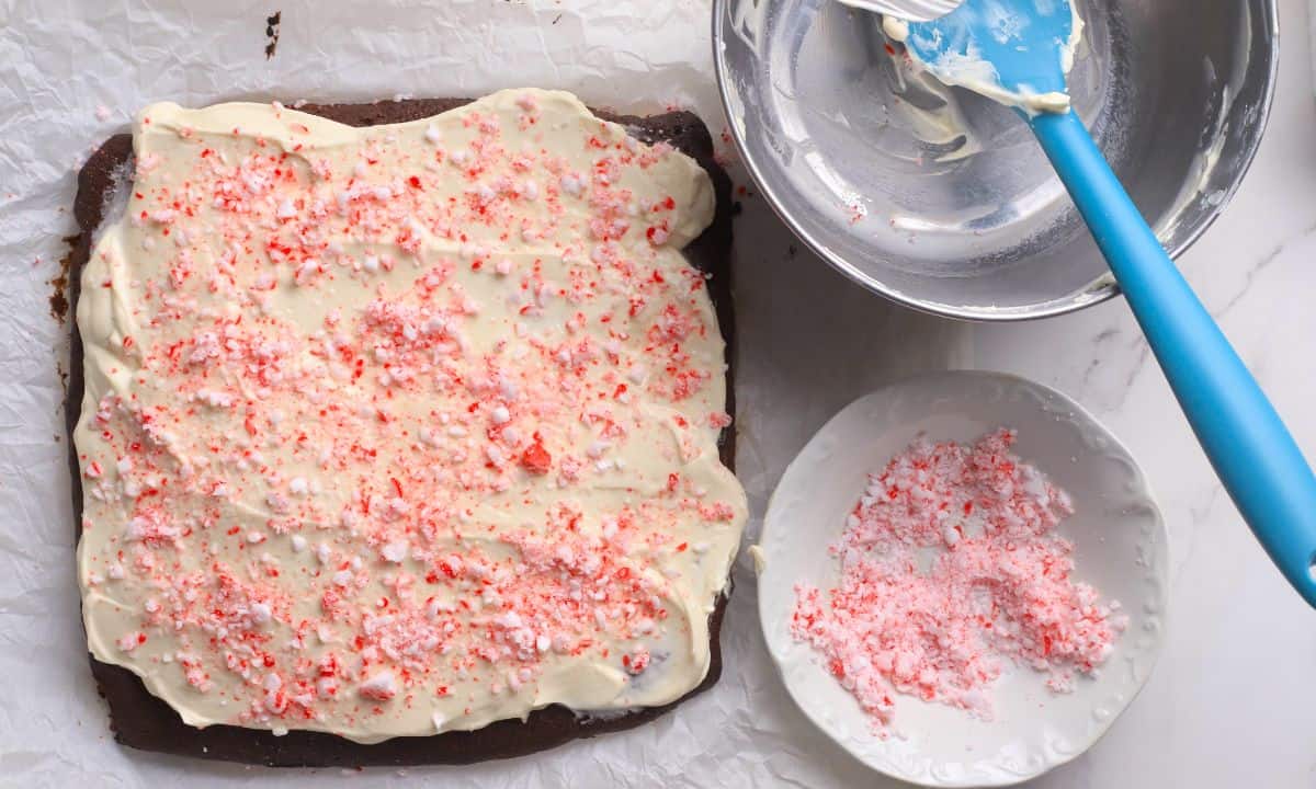 Low carb candy cane chocolate cake squares on a parchment paper next to a bowl with crushed peppermint candy and a bowl with a rubber spatula.