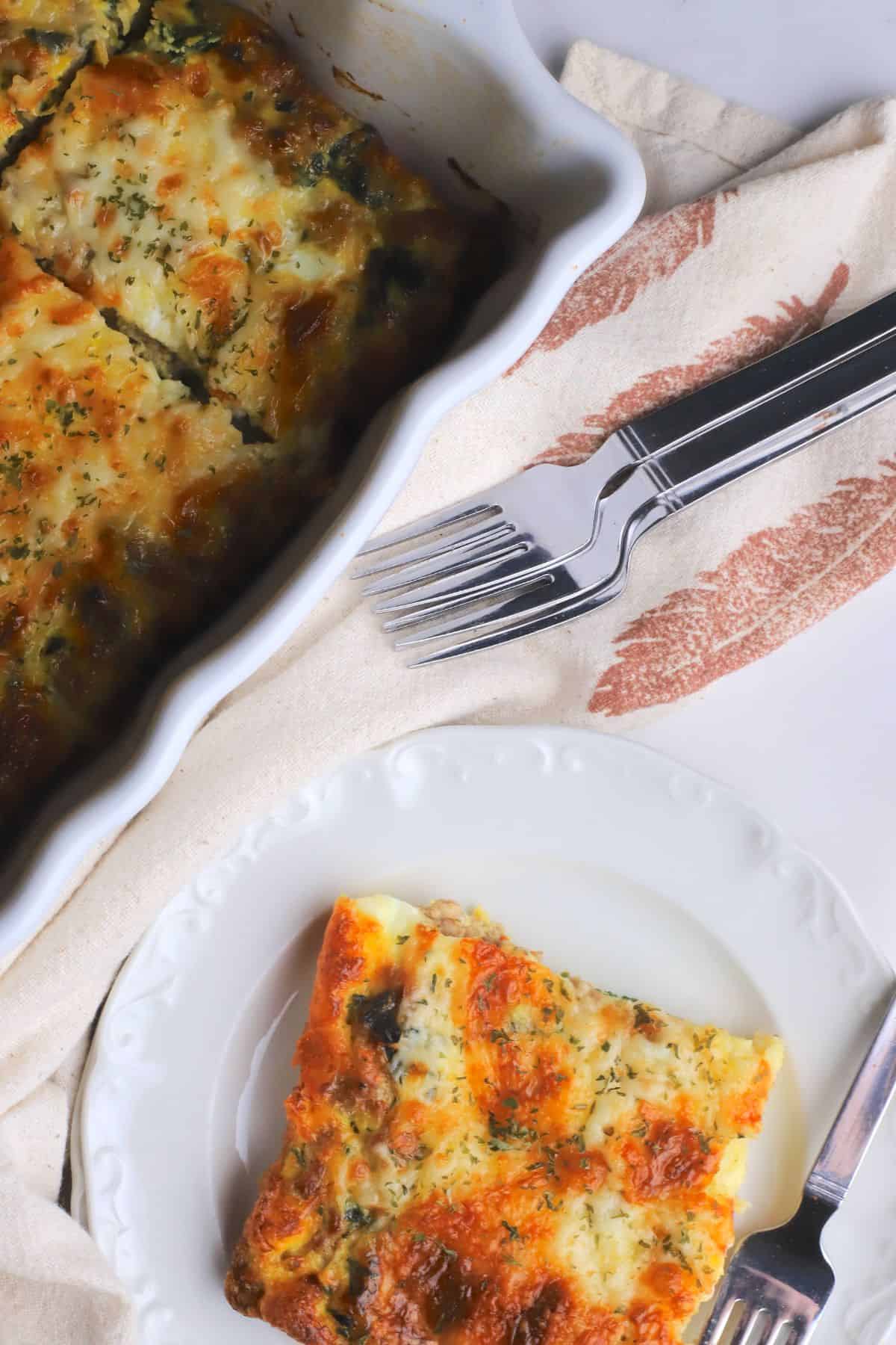 Low carb frittata slice on a side plate with a fork next to the casserole with more frittata.