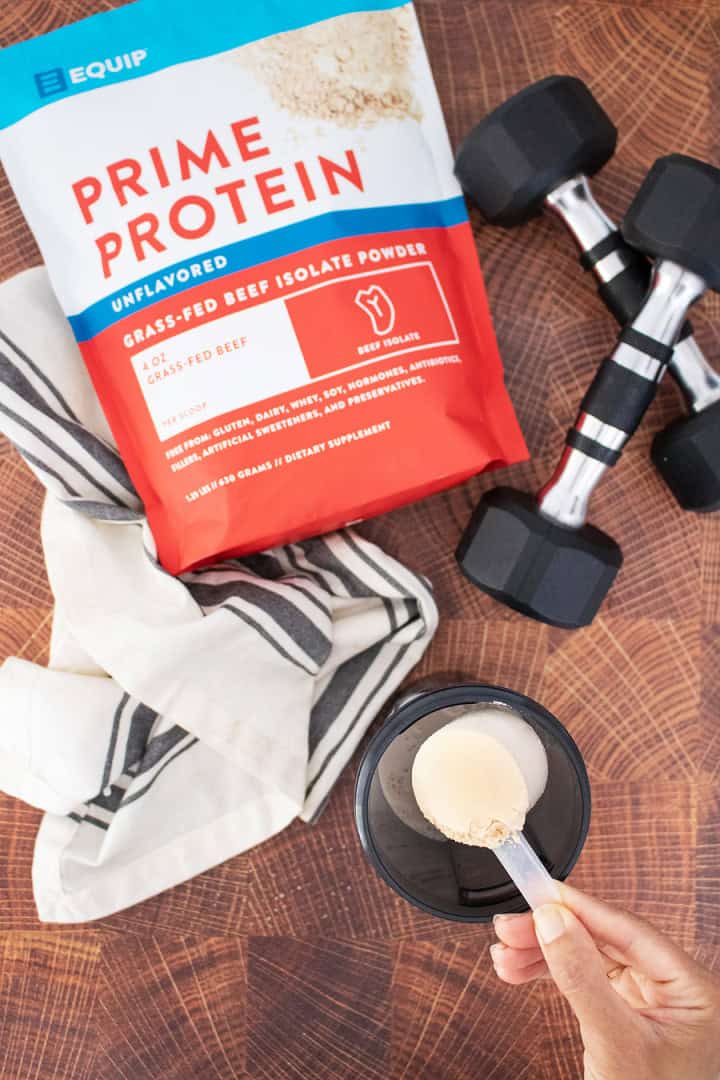 Product image for prime protein with dumbells and a cup with a scoop next to it under a towel.
