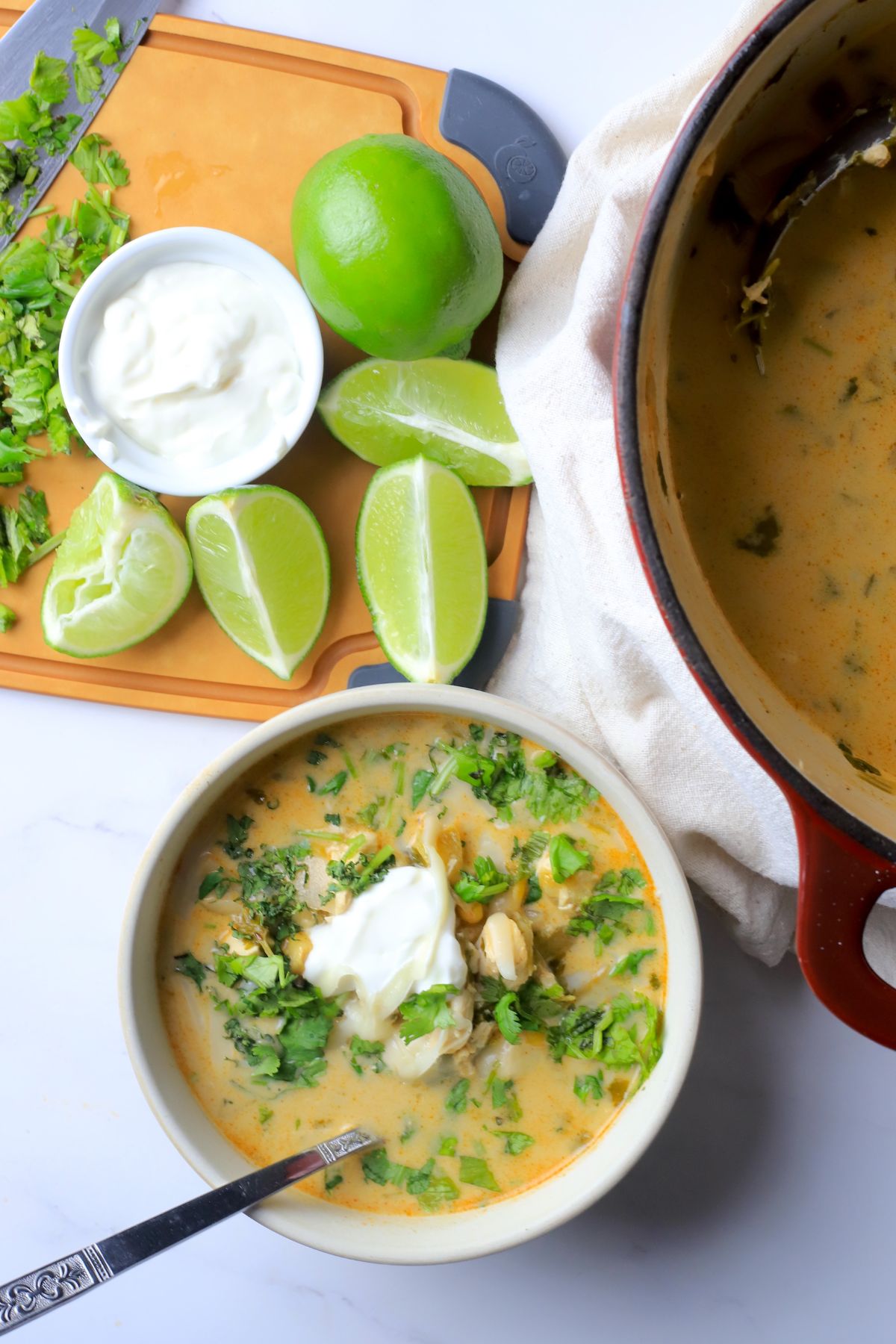Low carb white chicken chili in a bowl topped with cilantro and sour cream next to some limes and a pot of the same.