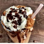 Large glass with sugar free cappuccino topped with whipping cream and chocolate with a plastic straw.
