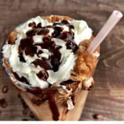 Large glass with sugar free cappuccino topped with whipping cream and chocolate with a plastic straw.