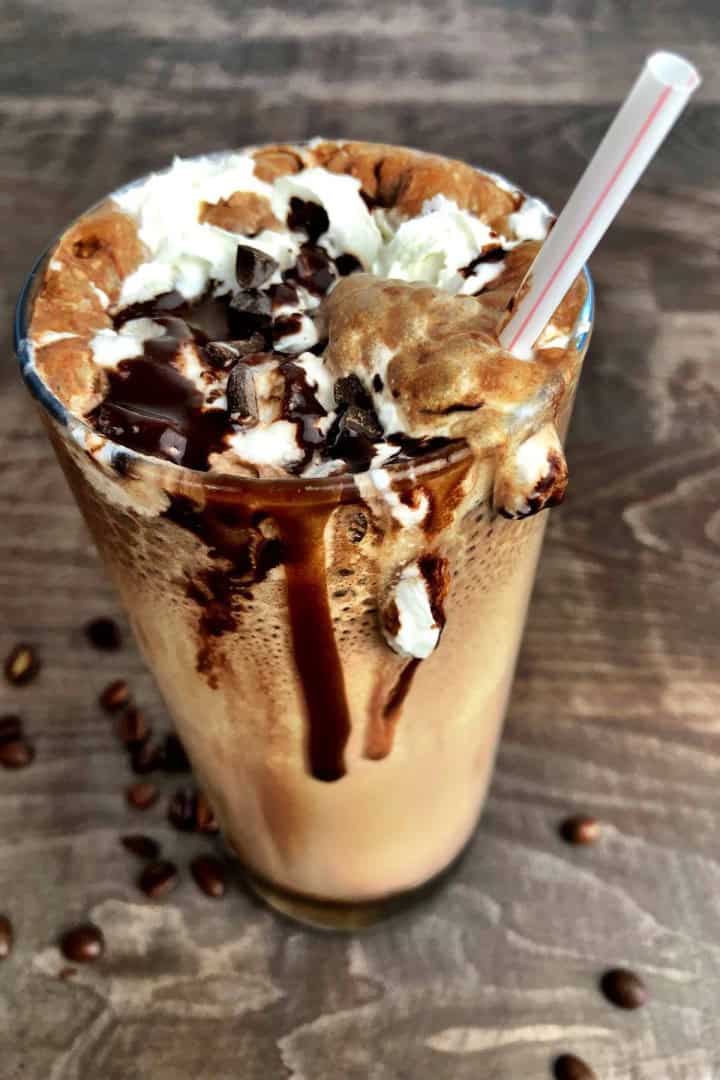 Large glass with sugar free cappuccino topped with whipping cream and chocolate with a plastic straw.