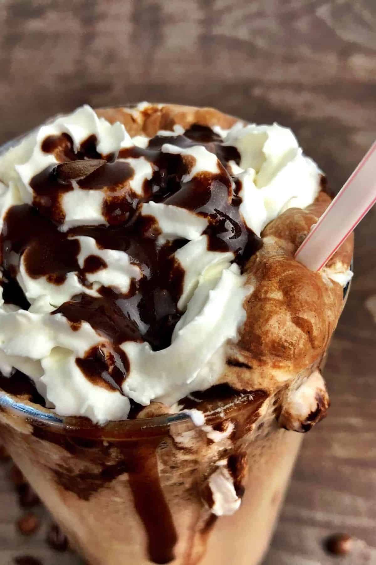 Large glass with sugar free cappuccino topped with whipping cream and chocolate with a plastic straw.
