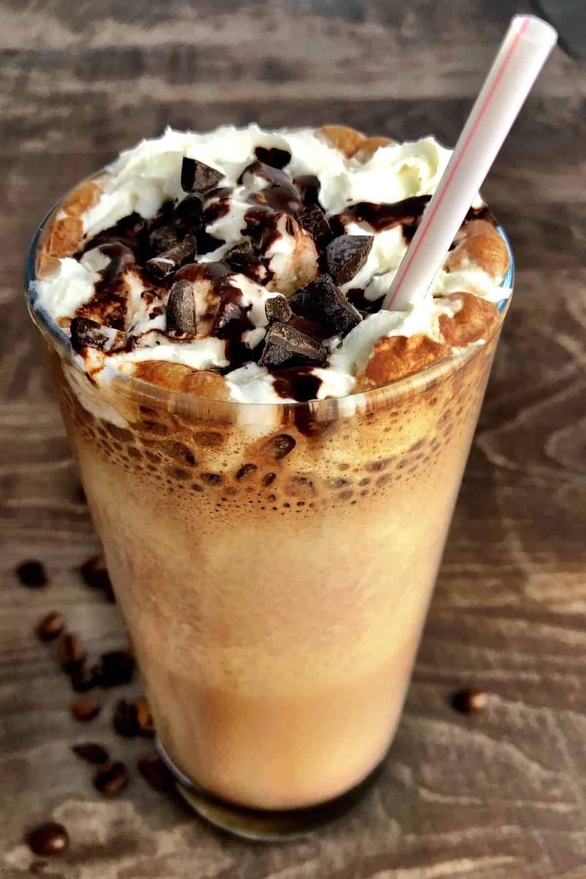 Large glass with sugar free cappuccino topped with whipping cream and chocolate with a plastic straw.
