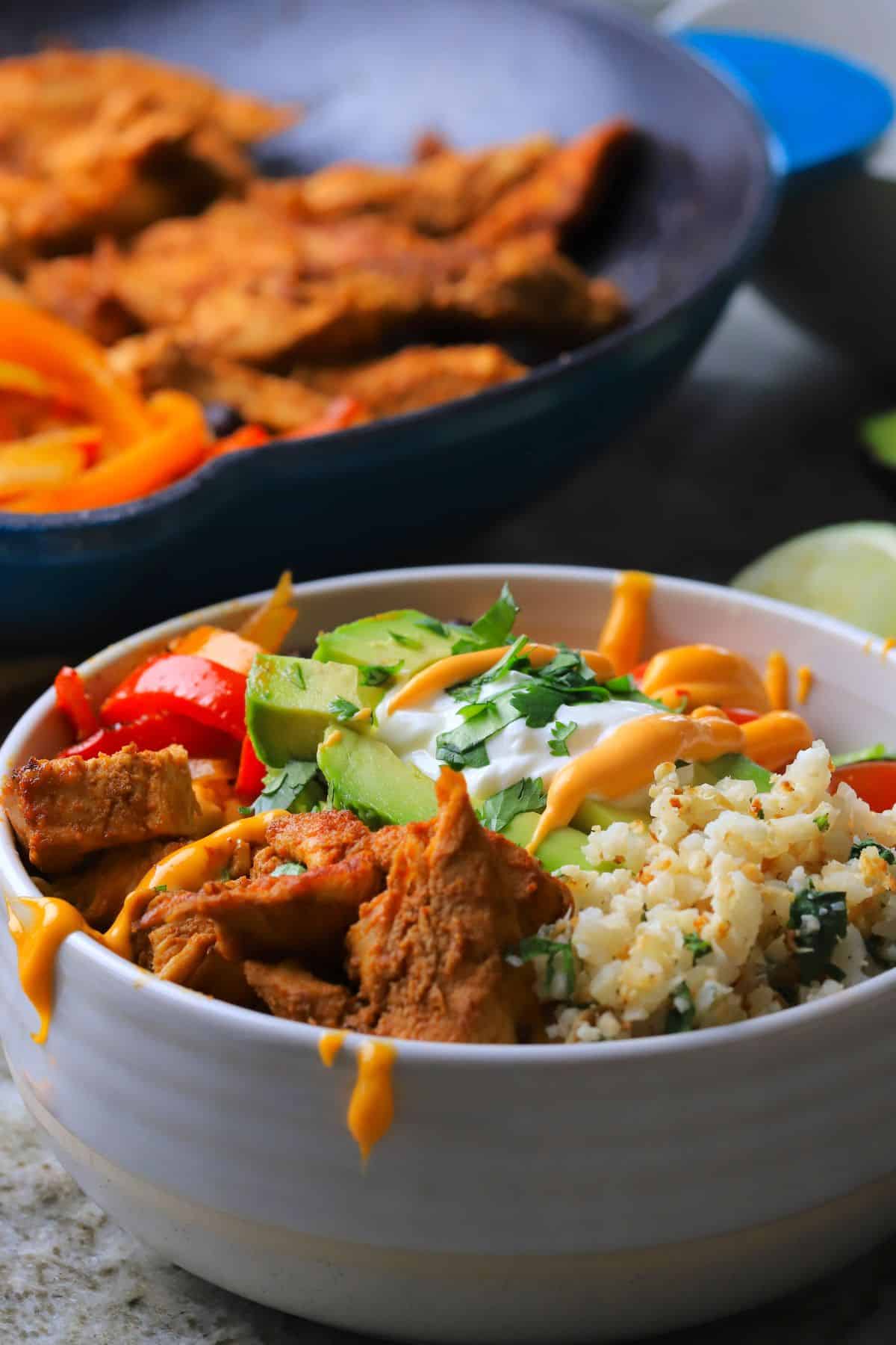 Low carb chicken fajita bowl filled with chicken, peppers, black beans, tomatoes, cauliflower rice and an avocado, next to a skillet.