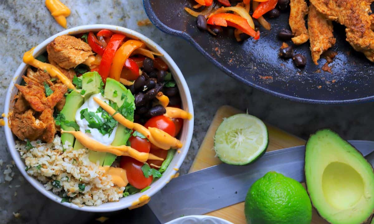 Low carb chicken fajita bowl filled with chicken, peppers, black beans, tomatoes, cauliflower rice and an avocado next to a skillet and limes and a half of an avocado.
