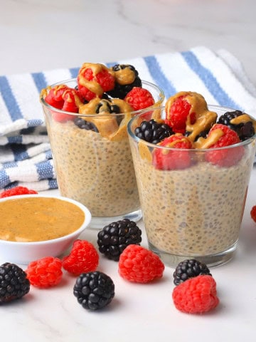 Two cups with low carb chia almond butter pudding topped with berries and almond butter with almond butter and berries on the side.